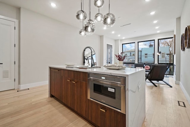 kitchen with an island with sink, stainless steel oven, pendant lighting, sink, and light hardwood / wood-style flooring