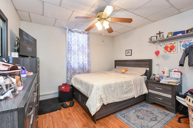 bedroom with a drop ceiling, a baseboard radiator, light wood-style flooring, and a ceiling fan