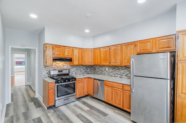 kitchen featuring light wood-style flooring, under cabinet range hood, stainless steel appliances, light stone countertops, and tasteful backsplash