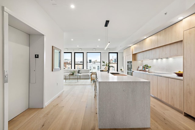 kitchen with light brown cabinetry, light wood-type flooring, sink, elevator, and an island with sink