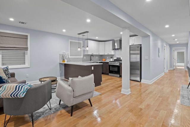 kitchen featuring white cabinets, wall chimney exhaust hood, appliances with stainless steel finishes, decorative light fixtures, and light hardwood / wood-style floors