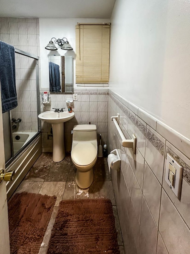 bathroom featuring tile walls, bath / shower combo with glass door, and toilet