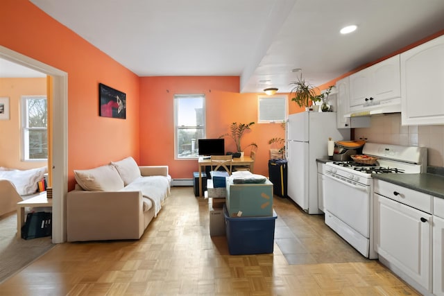 kitchen with dark countertops, baseboard heating, white cabinets, under cabinet range hood, and white appliances