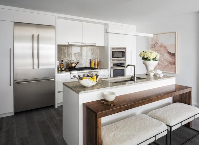 kitchen featuring white cabinets, a sink, built in appliances, under cabinet range hood, and dark wood-style floors