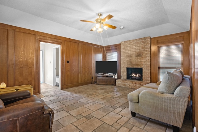 living room featuring wood walls, a textured ceiling, a brick fireplace, a raised ceiling, and ceiling fan