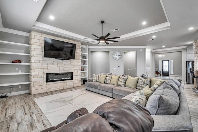 living room featuring crown molding, a tray ceiling, a stone fireplace, and built in shelves