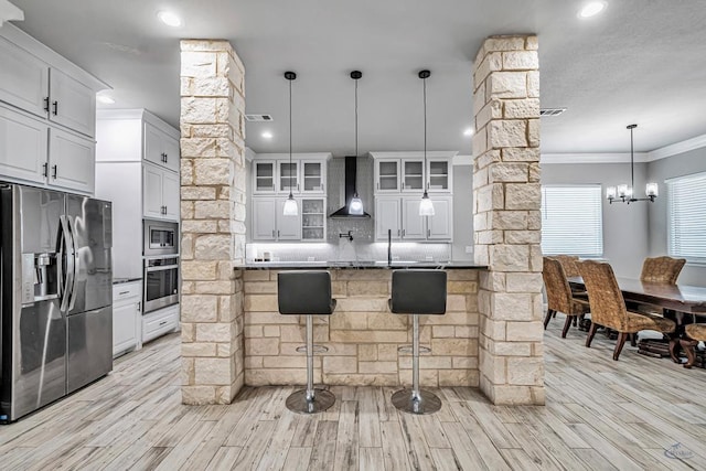 kitchen with decorative columns, white cabinets, a kitchen breakfast bar, stainless steel appliances, and wall chimney range hood