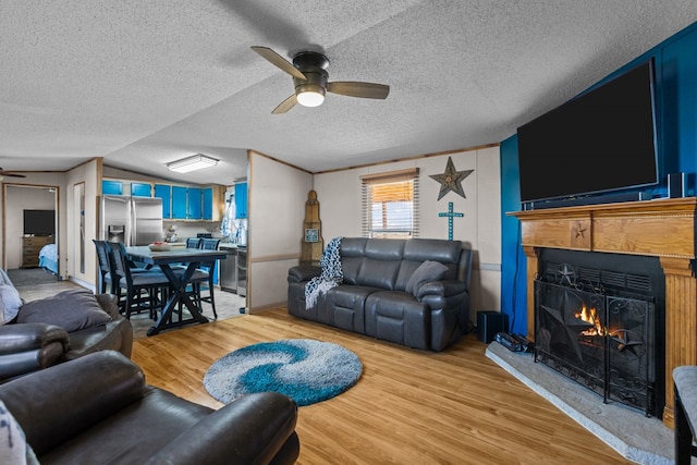 living area featuring light wood finished floors, ceiling fan, a lit fireplace, vaulted ceiling, and a textured ceiling