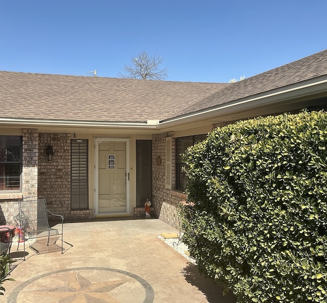 view of exterior entry featuring a patio, brick siding, and a shingled roof