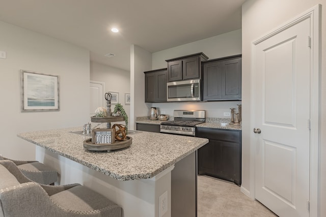 kitchen featuring dark brown cabinetry, stainless steel appliances, light stone counters, an island with sink, and a breakfast bar