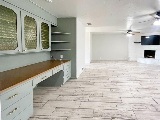 unfurnished office featuring a ceiling fan, visible vents, wood tiled floor, built in study area, and a brick fireplace