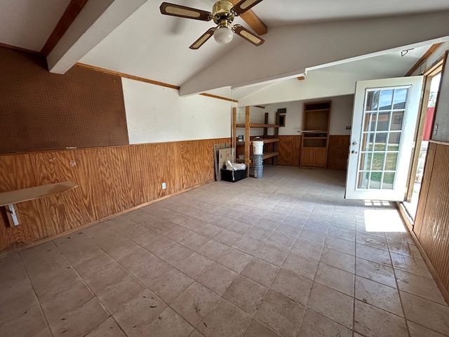 spare room featuring ceiling fan, wainscoting, wood walls, lofted ceiling with beams, and stone tile floors