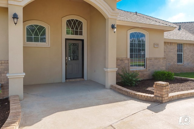 view of exterior entry featuring stucco siding, brick siding, and a shingled roof