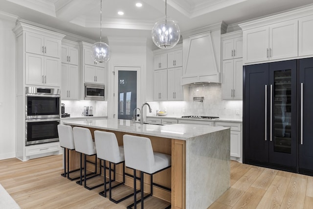 kitchen featuring custom exhaust hood, white cabinets, a center island with sink, built in appliances, and light wood-type flooring