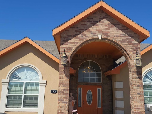 view of exterior entry with roof with shingles, stucco siding, and brick siding