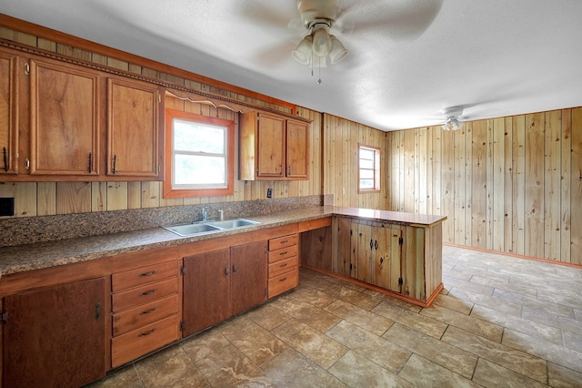 kitchen with a sink, a ceiling fan, brown cabinets, a peninsula, and wood walls