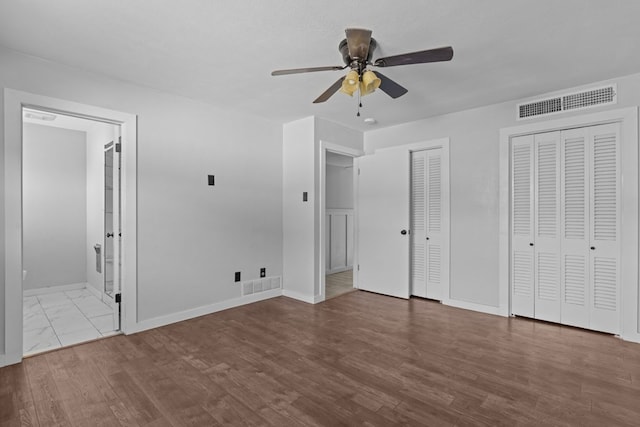 unfurnished bedroom featuring baseboards, two closets, ensuite bath, wood finished floors, and ceiling fan