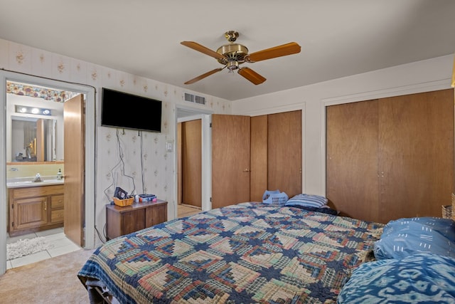 bedroom featuring two closets, wallpapered walls, ensuite bath, a ceiling fan, and a sink