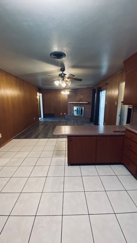 kitchen featuring wooden walls, ceiling fan, dark countertops, a peninsula, and a brick fireplace