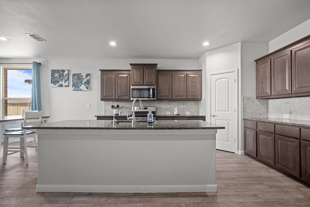 kitchen with stainless steel microwave, a sink, dark stone countertops, a center island with sink, and tasteful backsplash