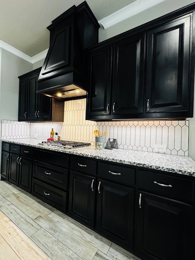 kitchen with dark cabinetry, light stone countertops, premium range hood, ornamental molding, and stainless steel gas stovetop