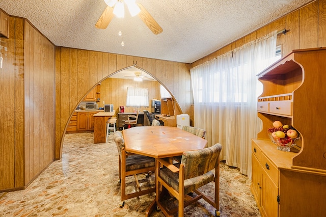 dining area with arched walkways, a textured ceiling, wood walls, and a ceiling fan