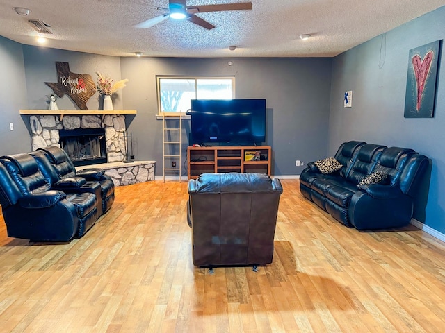 living room featuring a textured ceiling, light hardwood / wood-style flooring, ceiling fan, and a stone fireplace