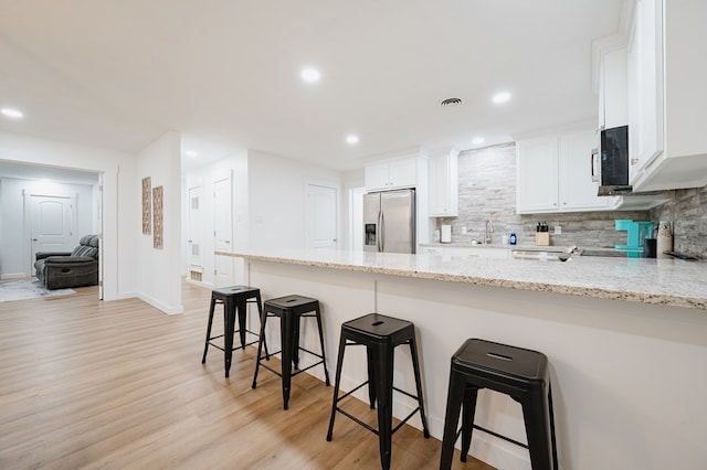 kitchen featuring decorative backsplash, stainless steel refrigerator with ice dispenser, light stone counters, a breakfast bar, and white cabinets