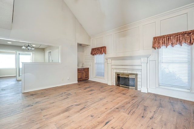 unfurnished living room featuring baseboards, light wood finished floors, high vaulted ceiling, a tile fireplace, and a chandelier