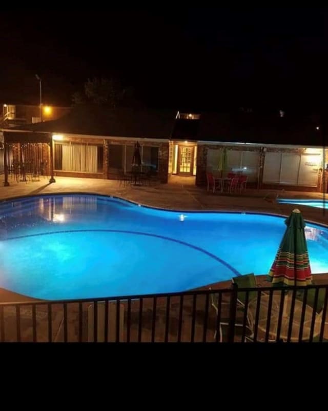 pool at night featuring a patio, fence, and a community pool