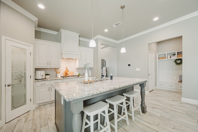 kitchen with decorative backsplash, custom range hood, light stone counters, light wood-type flooring, and stainless steel gas stovetop