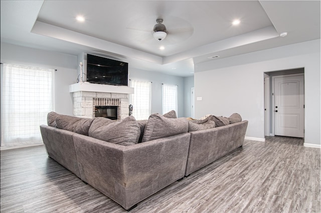 living room with a tray ceiling, a stone fireplace, ceiling fan, and wood-type flooring