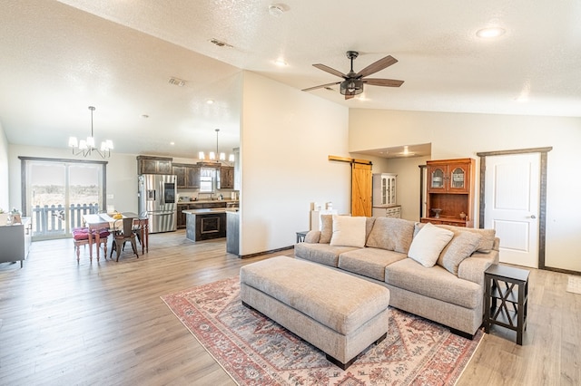 living room featuring vaulted ceiling, ceiling fan with notable chandelier, a barn door, a textured ceiling, and light hardwood / wood-style flooring