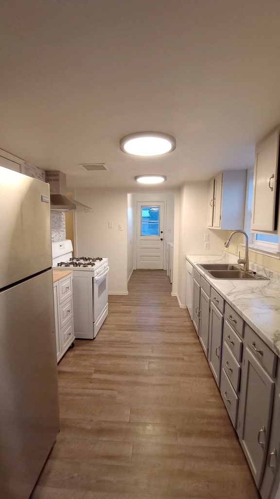 kitchen featuring wall chimney range hood, sink, light wood-type flooring, white gas range, and stainless steel refrigerator