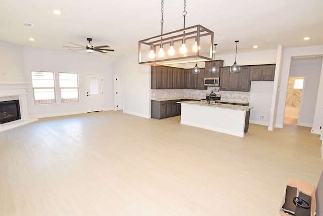 kitchen featuring pendant lighting, a fireplace, decorative backsplash, a kitchen island with sink, and dark brown cabinetry