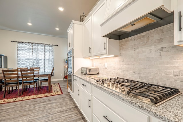 kitchen with tasteful backsplash, appliances with stainless steel finishes, white cabinets, custom range hood, and light wood-type flooring