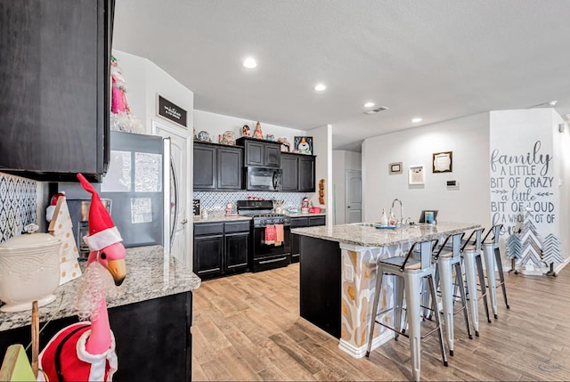 kitchen with black gas range, light stone countertops, light hardwood / wood-style flooring, a kitchen island with sink, and a breakfast bar