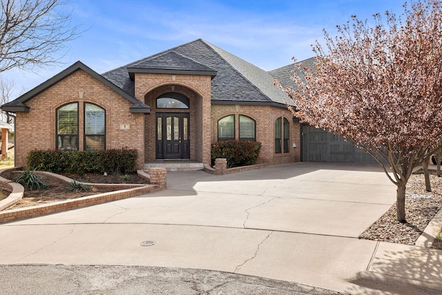 french country inspired facade with concrete driveway, brick siding, and a shingled roof