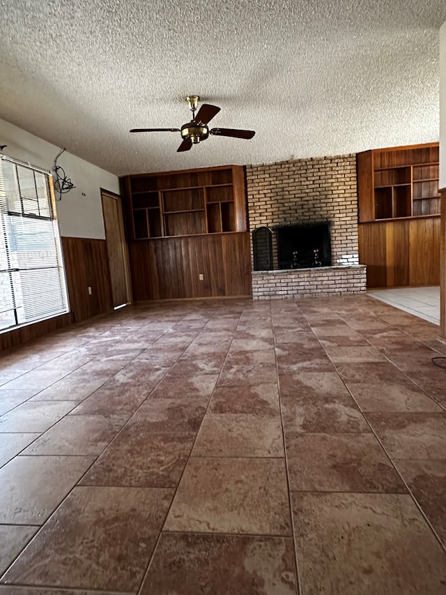 unfurnished living room featuring wooden walls, ceiling fan, a brick fireplace, a textured ceiling, and built in shelves