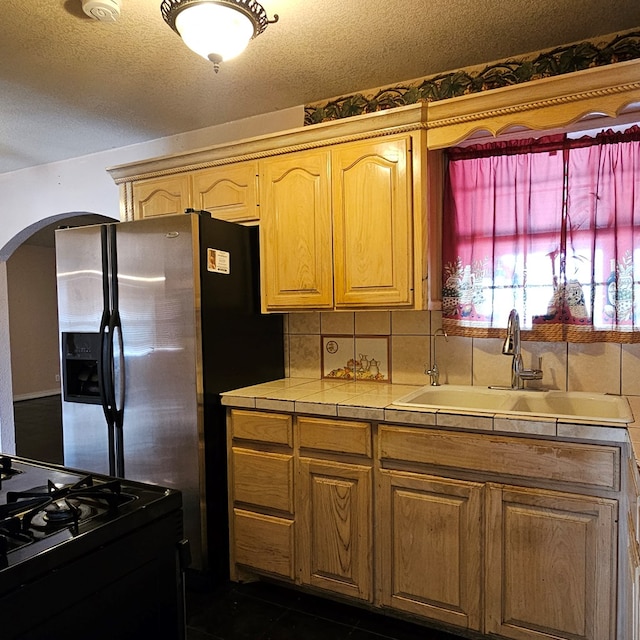 kitchen featuring arched walkways, tile countertops, decorative backsplash, black range with gas cooktop, and a sink