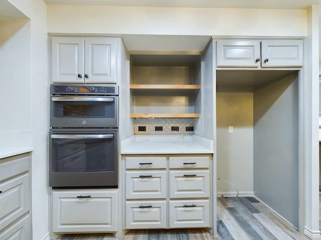 kitchen with white cabinetry, stainless steel double oven, tasteful backsplash, light stone counters, and light hardwood / wood-style flooring