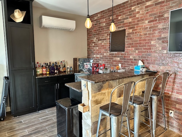 bar with dark stone countertops, hanging light fixtures, a wall unit AC, light hardwood / wood-style floors, and brick wall