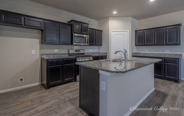 kitchen featuring sink, appliances with stainless steel finishes, a kitchen island with sink, dark hardwood / wood-style floors, and dark stone counters