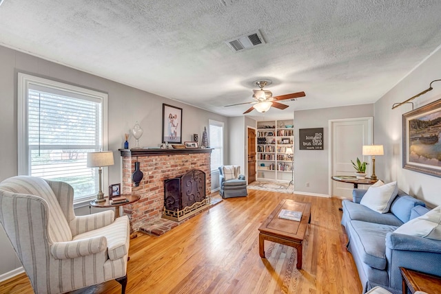 living area featuring visible vents, a textured ceiling, light wood-type flooring, a brick fireplace, and built in shelves