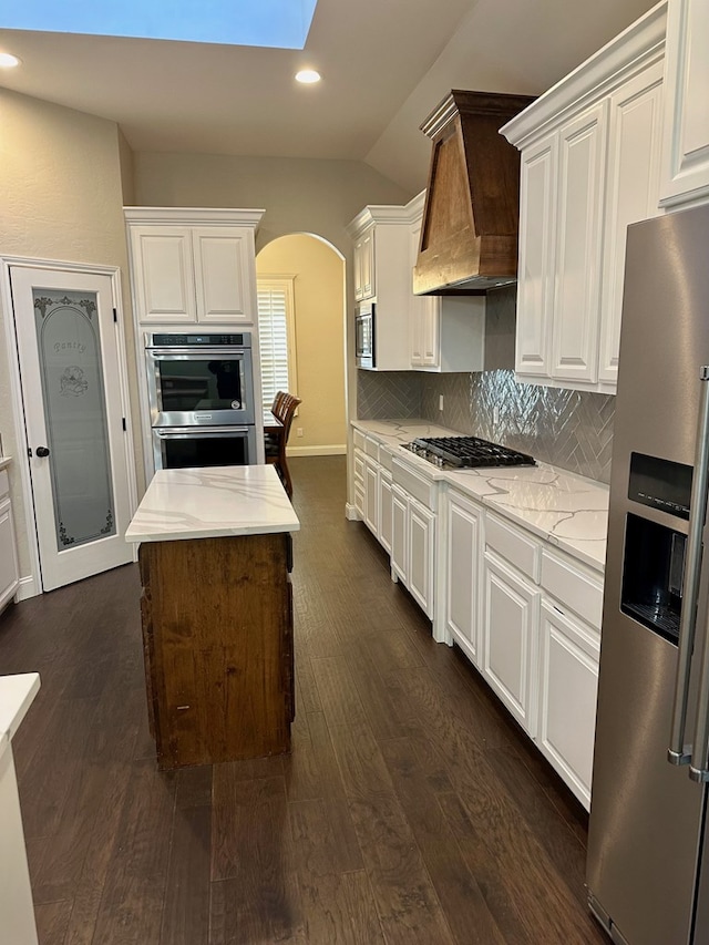 kitchen with custom exhaust hood, vaulted ceiling with skylight, arched walkways, appliances with stainless steel finishes, and backsplash