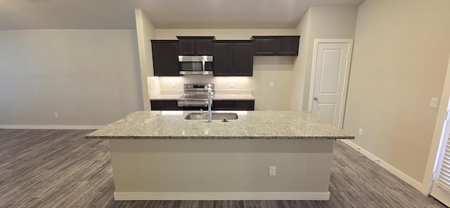 kitchen featuring light stone counters, baseboards, an island with sink, appliances with stainless steel finishes, and dark cabinets