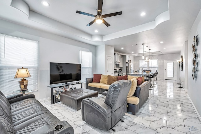 living room with a tray ceiling, marble finish floor, recessed lighting, baseboards, and ceiling fan with notable chandelier