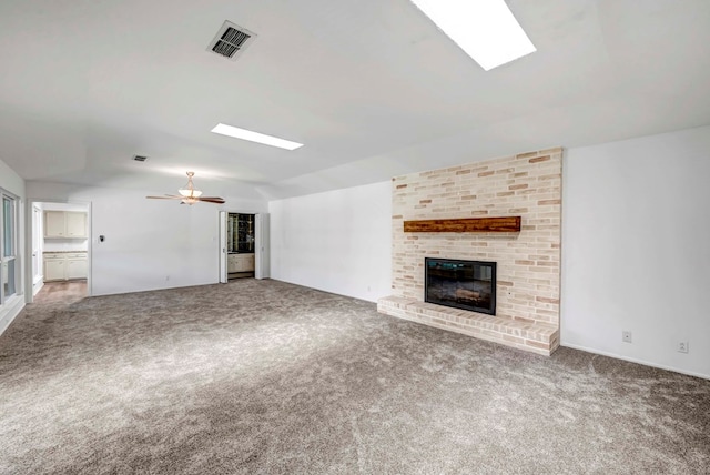 unfurnished living room with a brick fireplace, ceiling fan, vaulted ceiling with skylight, and carpet