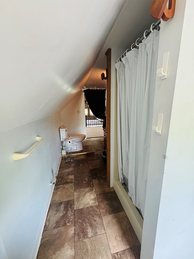 bathroom with vaulted ceiling, stone finish flooring, baseboards, toilet, and curtained shower