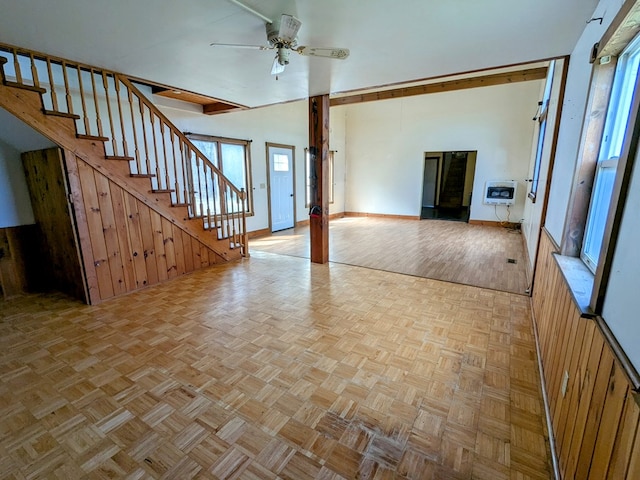 unfurnished living room featuring baseboards, ceiling fan, heating unit, wooden walls, and stairway